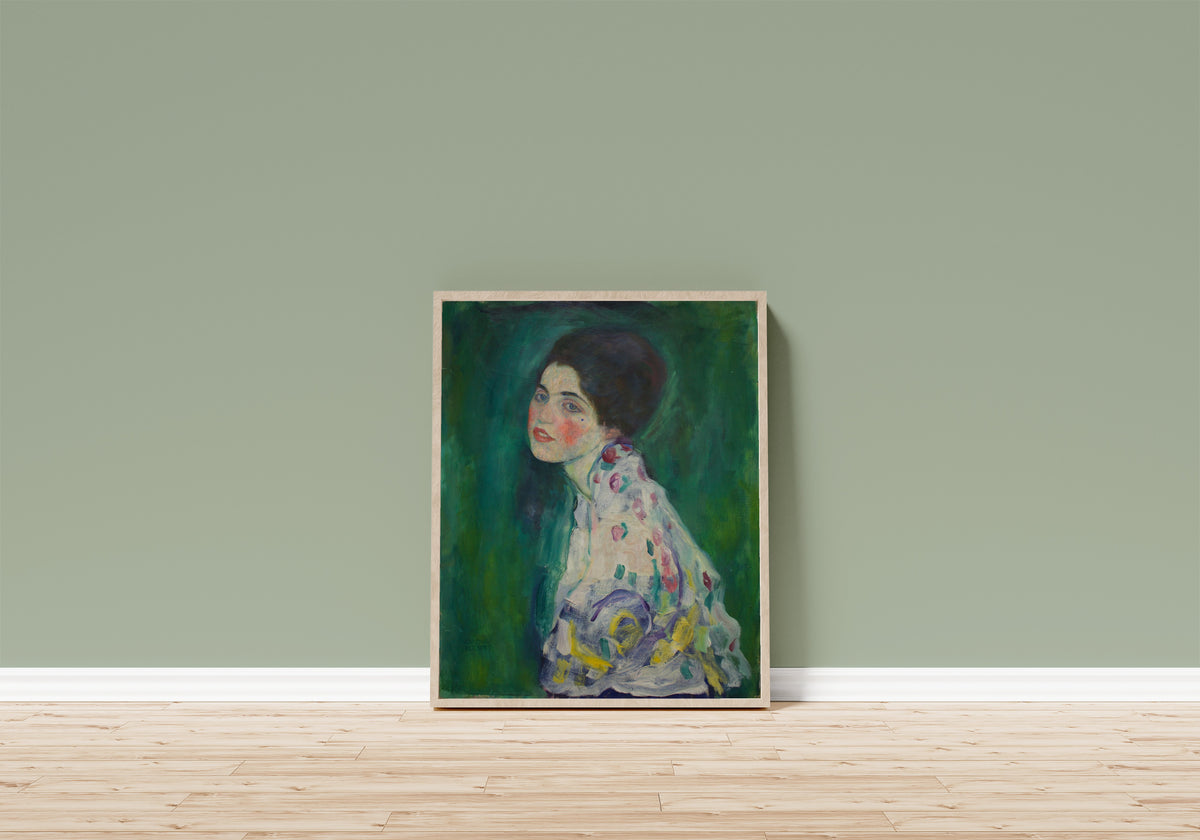 Framed art print of Portrait of a Lady by Gustav Klimt, in a light wood frame leaning on a sage green wall above a wooden floor