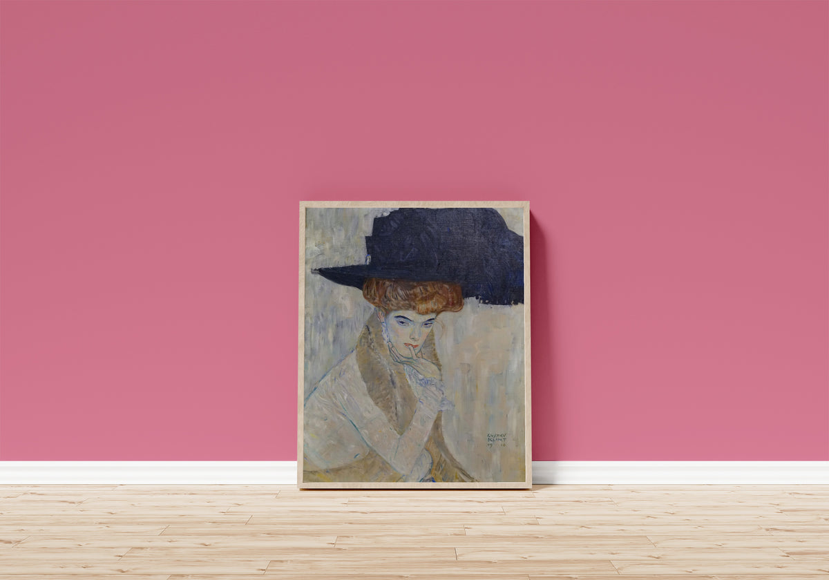 Framed art print of The Black Feathered Hat by Gustav Klimt, in a light wood frame leaning on a pink wall above a wooden floor
