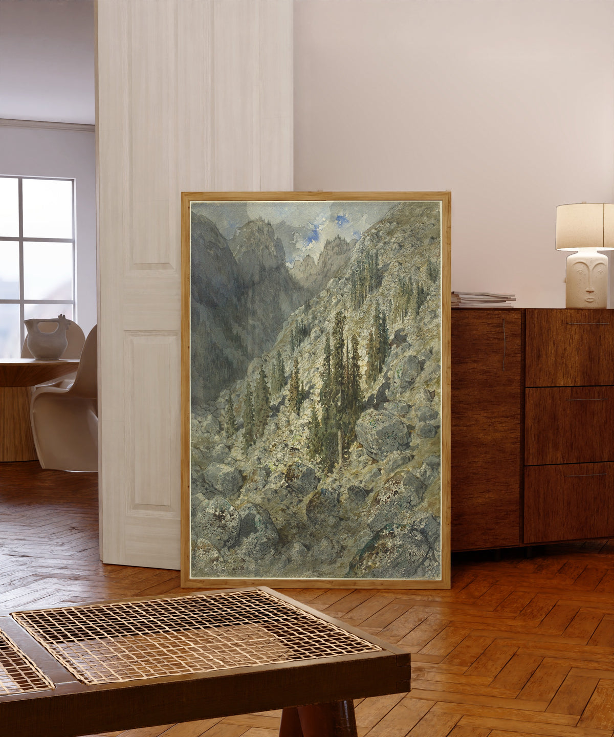 Framed art print of An Alpine Valley with Trees and Boulders by Gustave Doré, in a light wood frame leaning in a warm room with a parquet floor