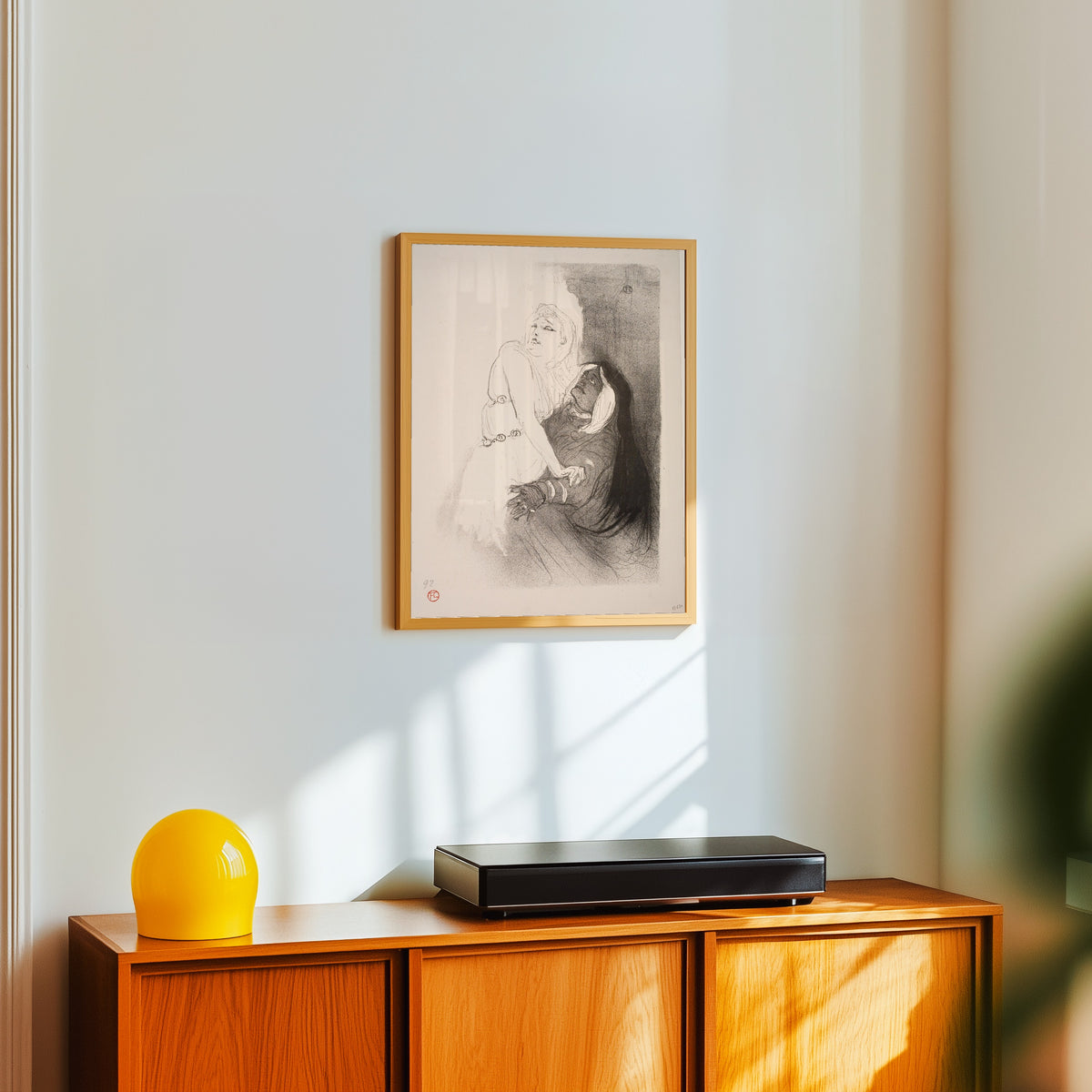 Framed art print of At the Renaissance: Sarah Bernhardt in Phèdre by Henri de Toulouse-Lautrec, in a light wood frame on a white wall above an orange sideboard