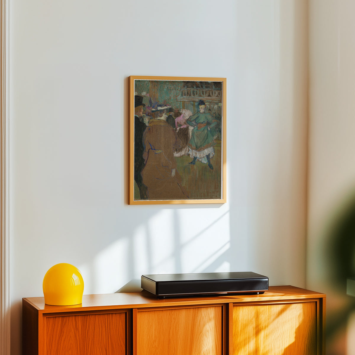 Framed art print of Quadrille at the Moulin Rouge by Henri de Toulouse-Lautrec, in a light wood frame on a white wall above an orange sideboard