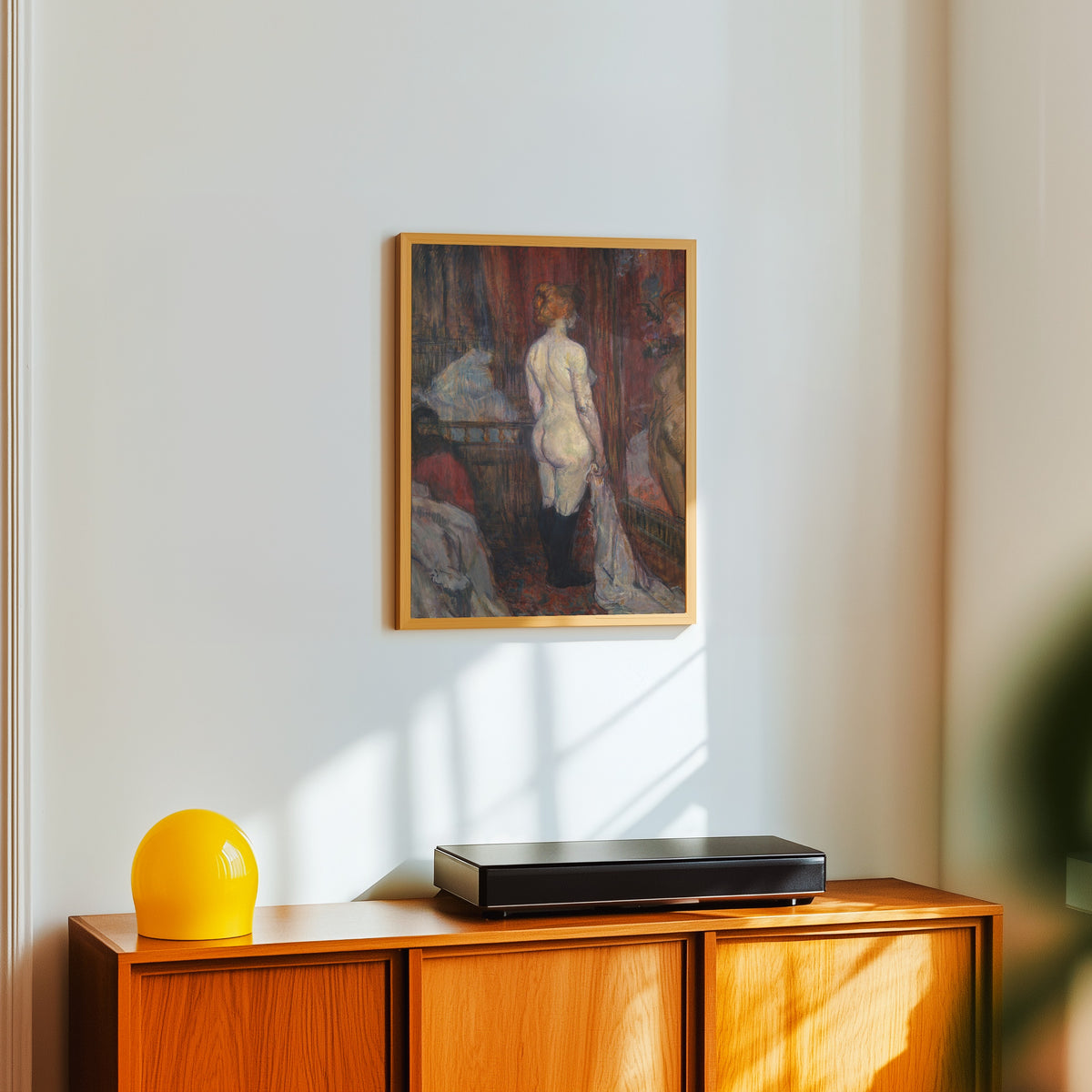 Framed art print of Woman before a Mirror by Henri de Toulouse-Lautrec, in a light wood frame on a white wall above an orange sideboard