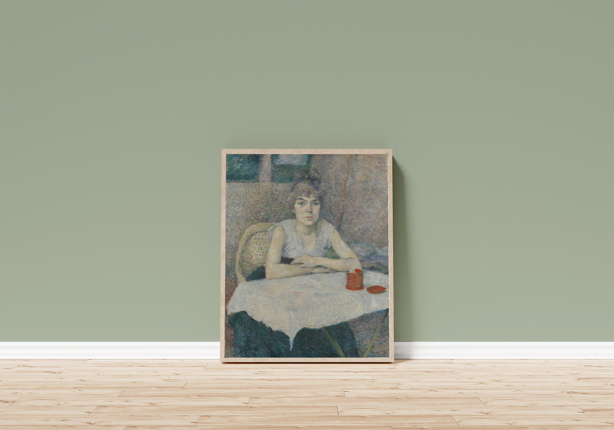 Framed art print of Young Woman at a Table, 'Poudre de… by Henri de Toulouse-Lautrec, in a light wood frame leaning on a sage green wall above a wooden floor