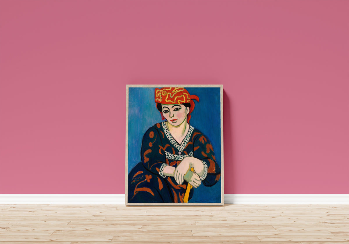 Framed art print of Red Madras Headdress (Le Madras rouge) by Henri Matisse, in a light wood frame leaning on a pink wall above a wooden floor