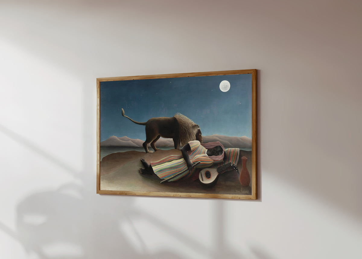 Framed art print of The Sleeping Gypsy by Henri Rousseau, in a light wood frame on a sunlit white wall, seen at a slight angle