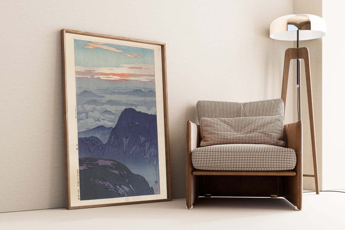 Framed art print of Sunrise on Eboshidake by Hiroshi Yoshida, leaning against a wall beside an armchair and a floor lamp