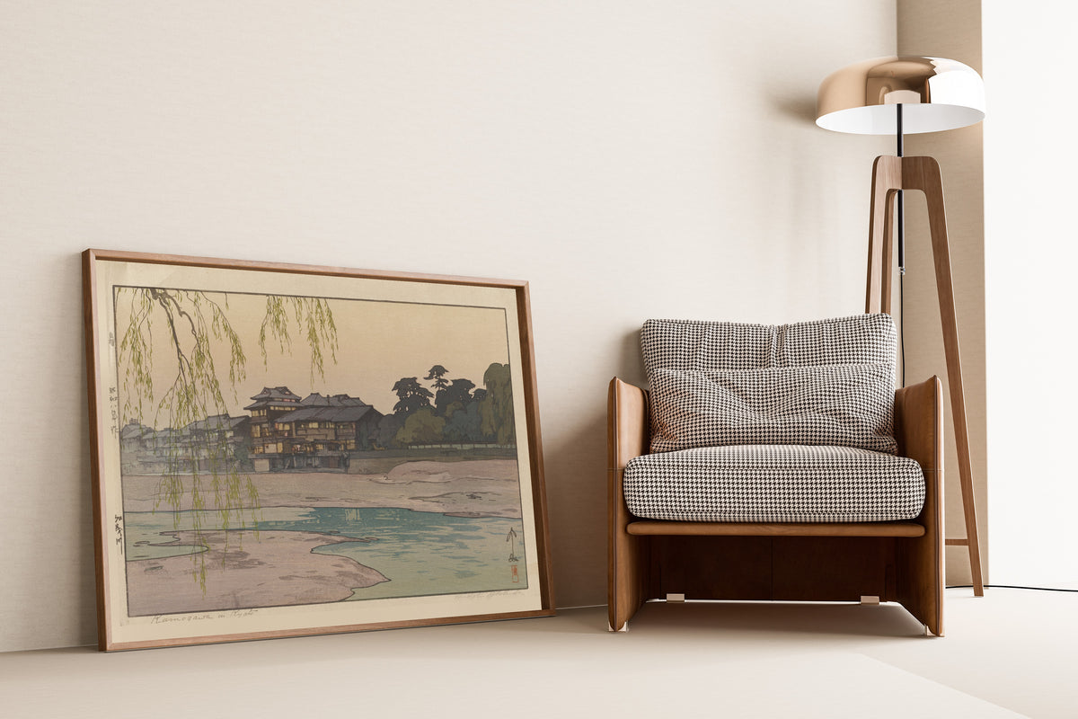 Framed art print of The Kamo River by Hiroshi Yoshida, leaning against a wall beside an armchair and a floor lamp