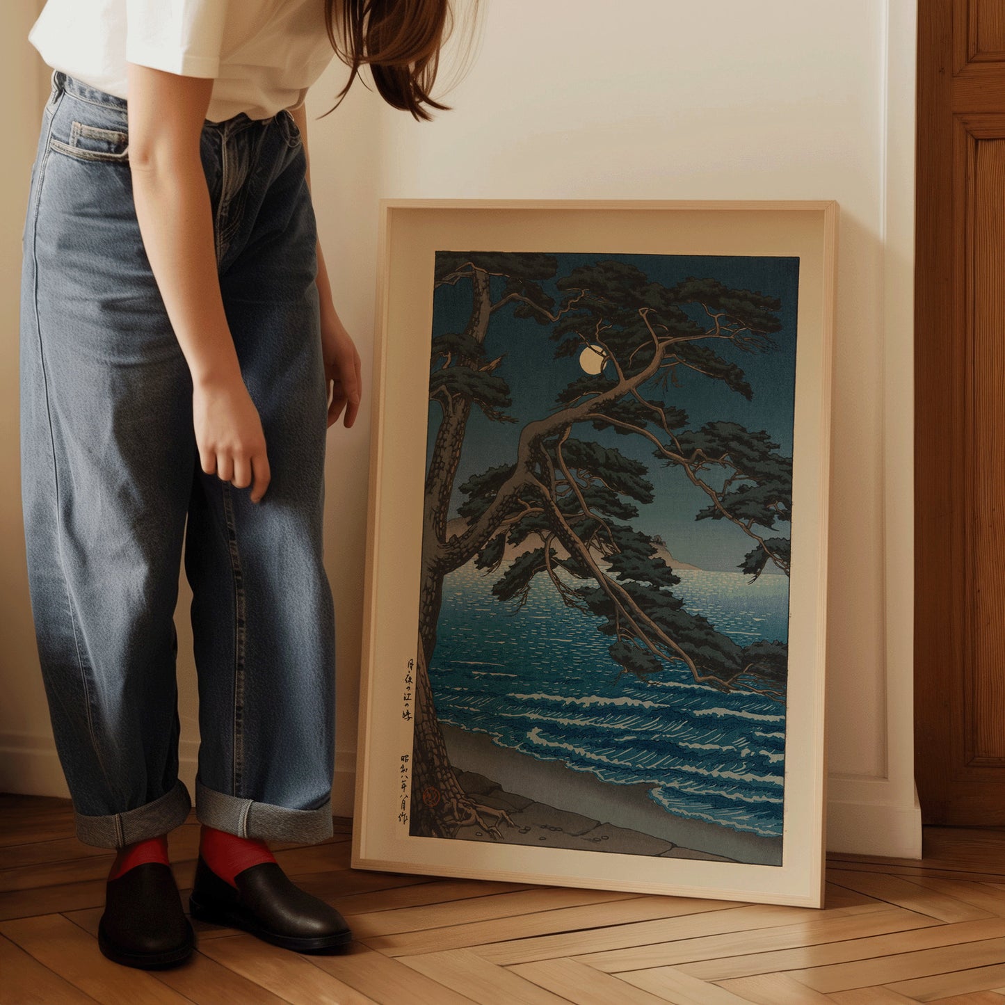 Person arranging framed Kawase Hasui's Moon at Enoshima Beach art print on floor, showcasing vintage Japanese woodblock style.