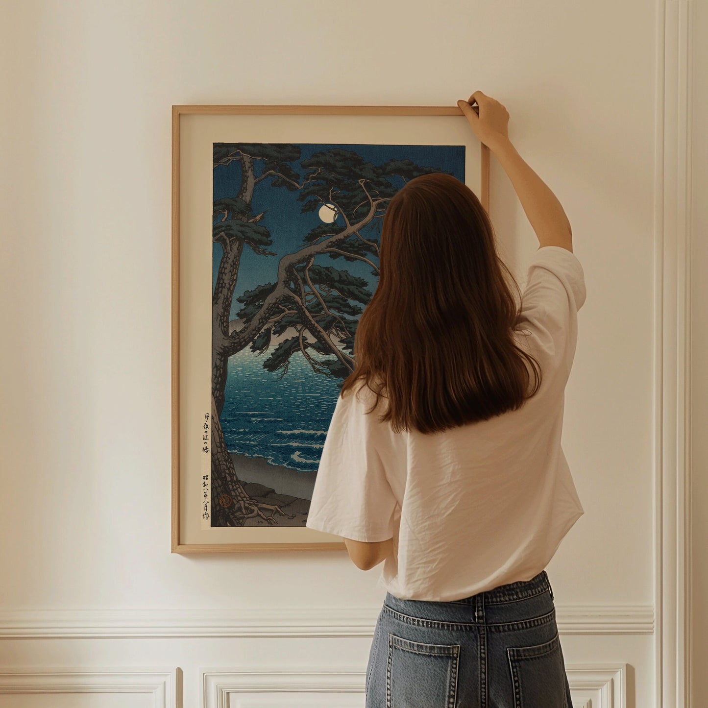 Woman hanging framed Kawase Hasui Moon at Enoshima Beach woodblock art on wall