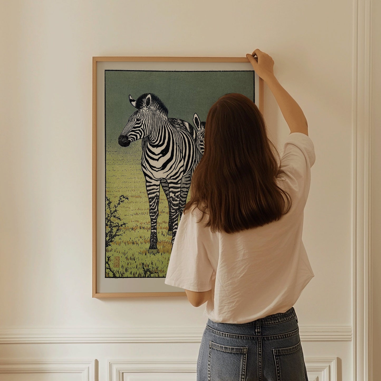 Woman hanging Toshi Yoshida's vintage Japanese zebra woodblock art in a yellow gold frame on a cream wall.