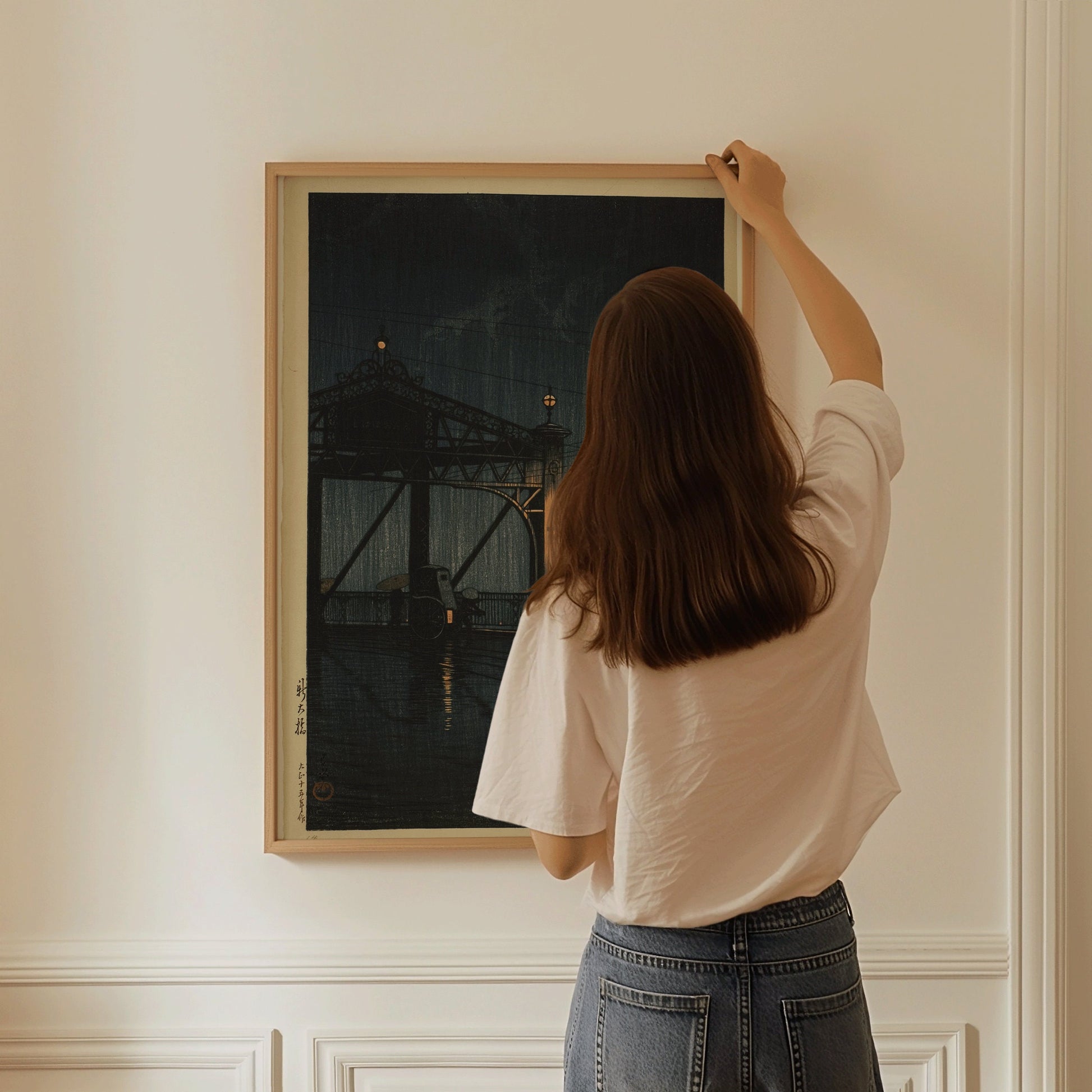 Woman hanging framed Kawase Hasui Shin Ohashi Bridge at Night woodblock print, showcasing vintage Japanese art.