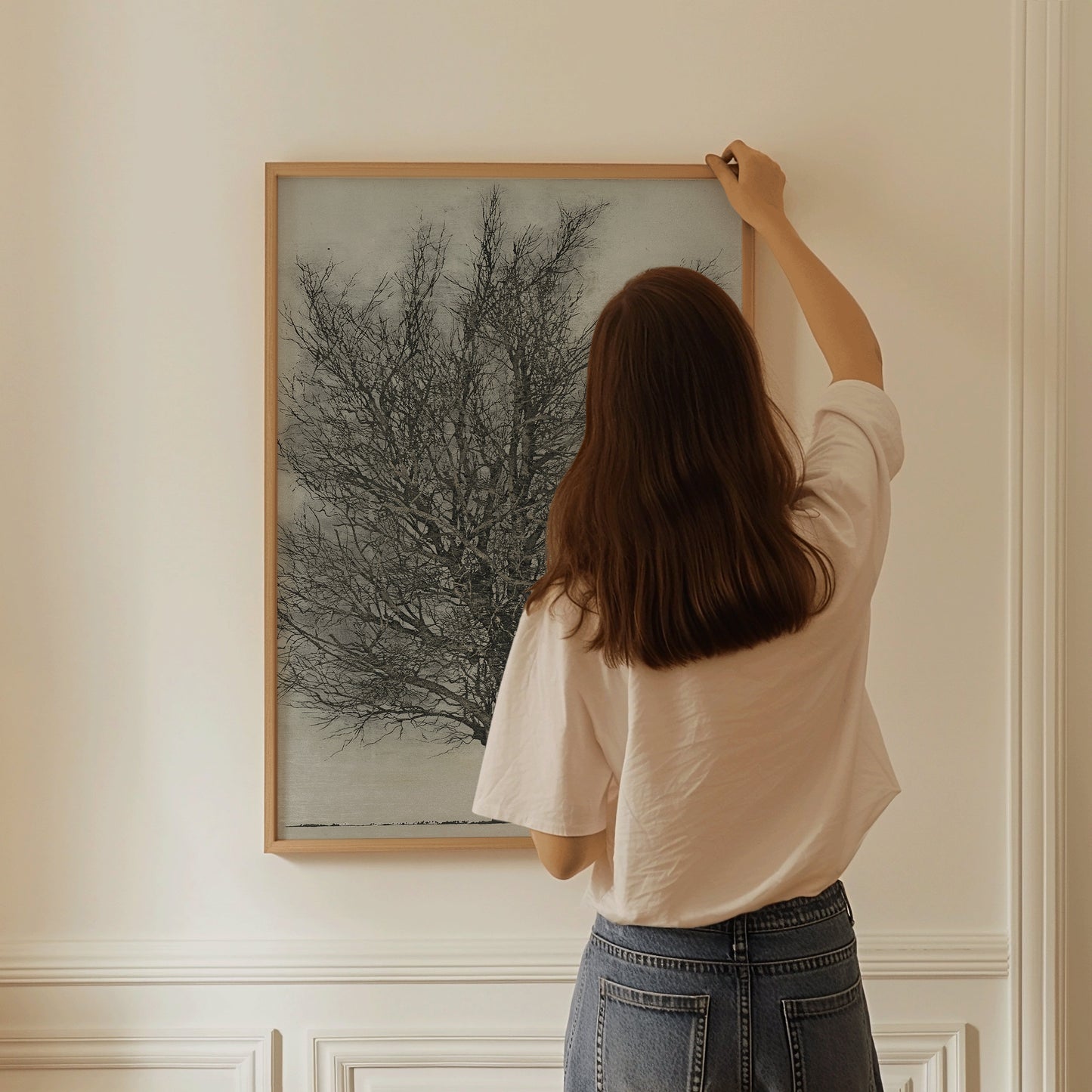 Woman hanging Joichi Hoshi's "The Black Tree" woodblock art in a nude oak frame on a white wall.