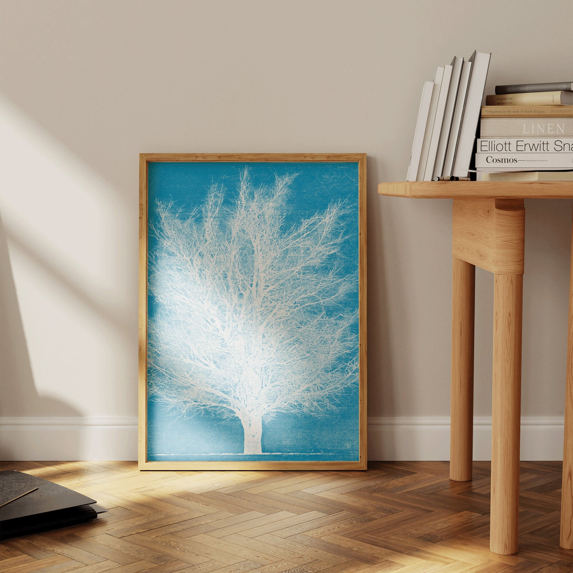 Vintage Japanese woodblock art print of a white tree on blue background, framed in nude oak, leaning against a wall near a wooden table.