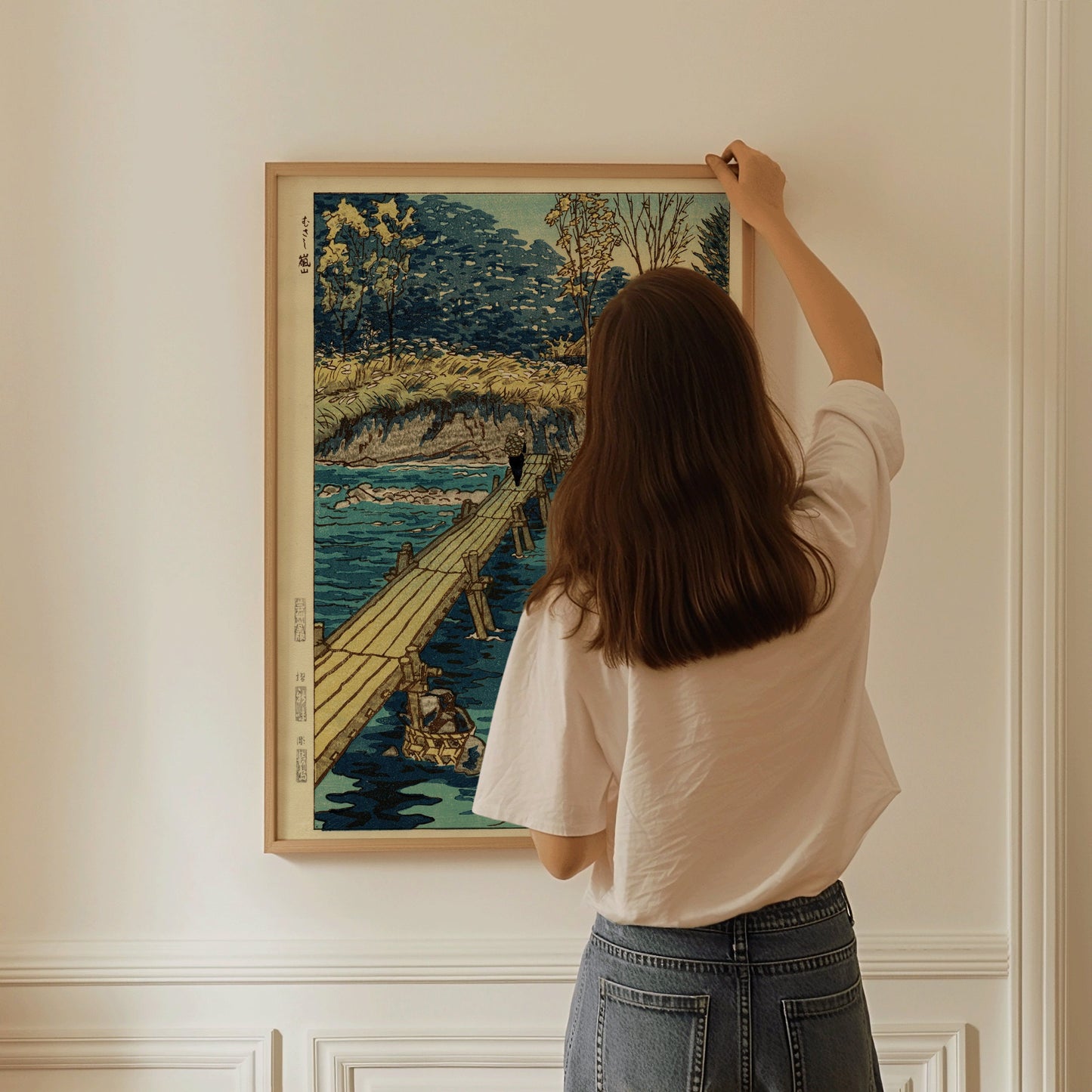 Woman hanging Shiro Kasamatsu's "Bridge at Musashi, Arashiyama" print in a natural wood frame on a white wall.