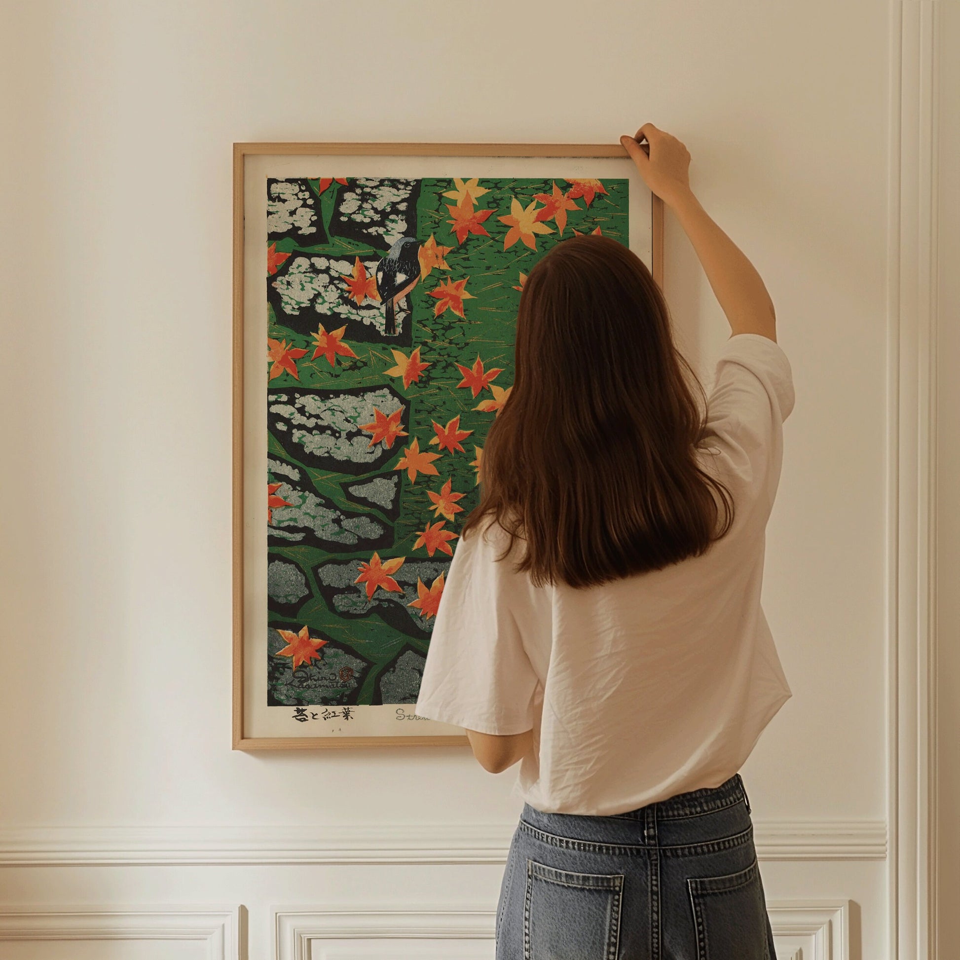 Woman hanging Shiro Kasamatsu's "Moss and Maple" framed print, highlighting Japanese Shin-Hanga art with vibrant autumn colors.