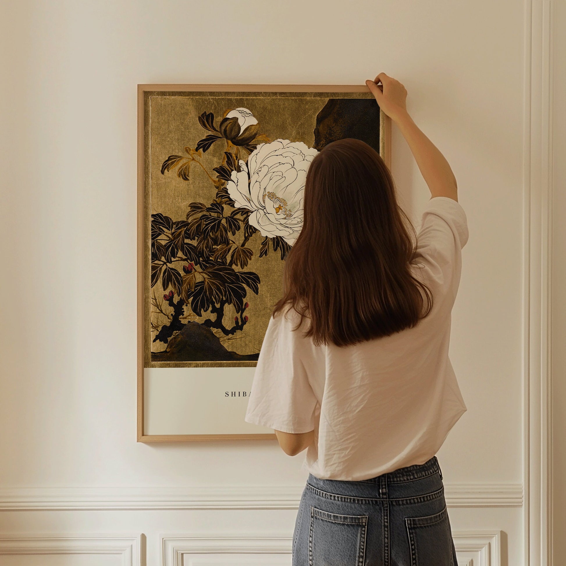 Woman hanging Shibata Zeshin's vintage Japanese woodblock art of peonies in a frame on a wall.