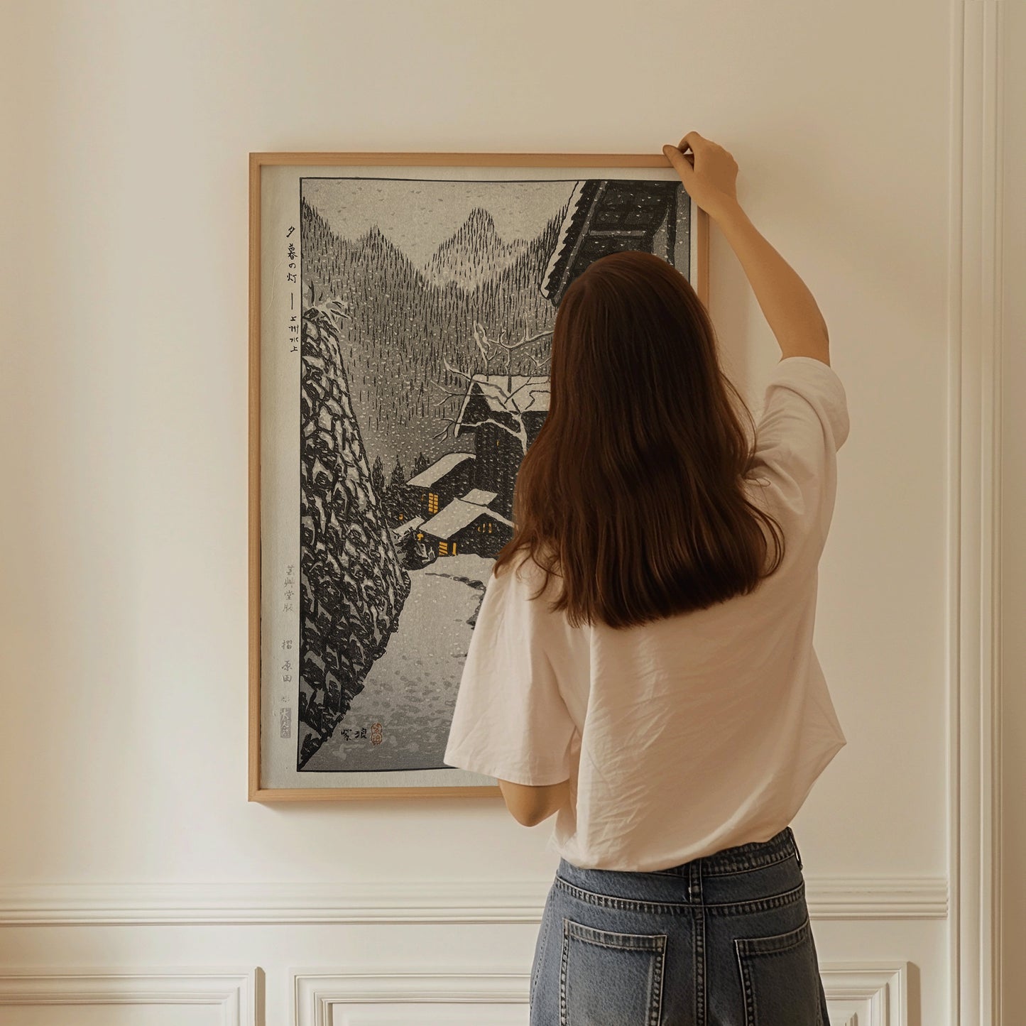 Woman hanging Shiro Kasamatsu's "Light in the Evening Snow Country" framed art on wall, showcasing neutral Japanese Shin-Hanga style.