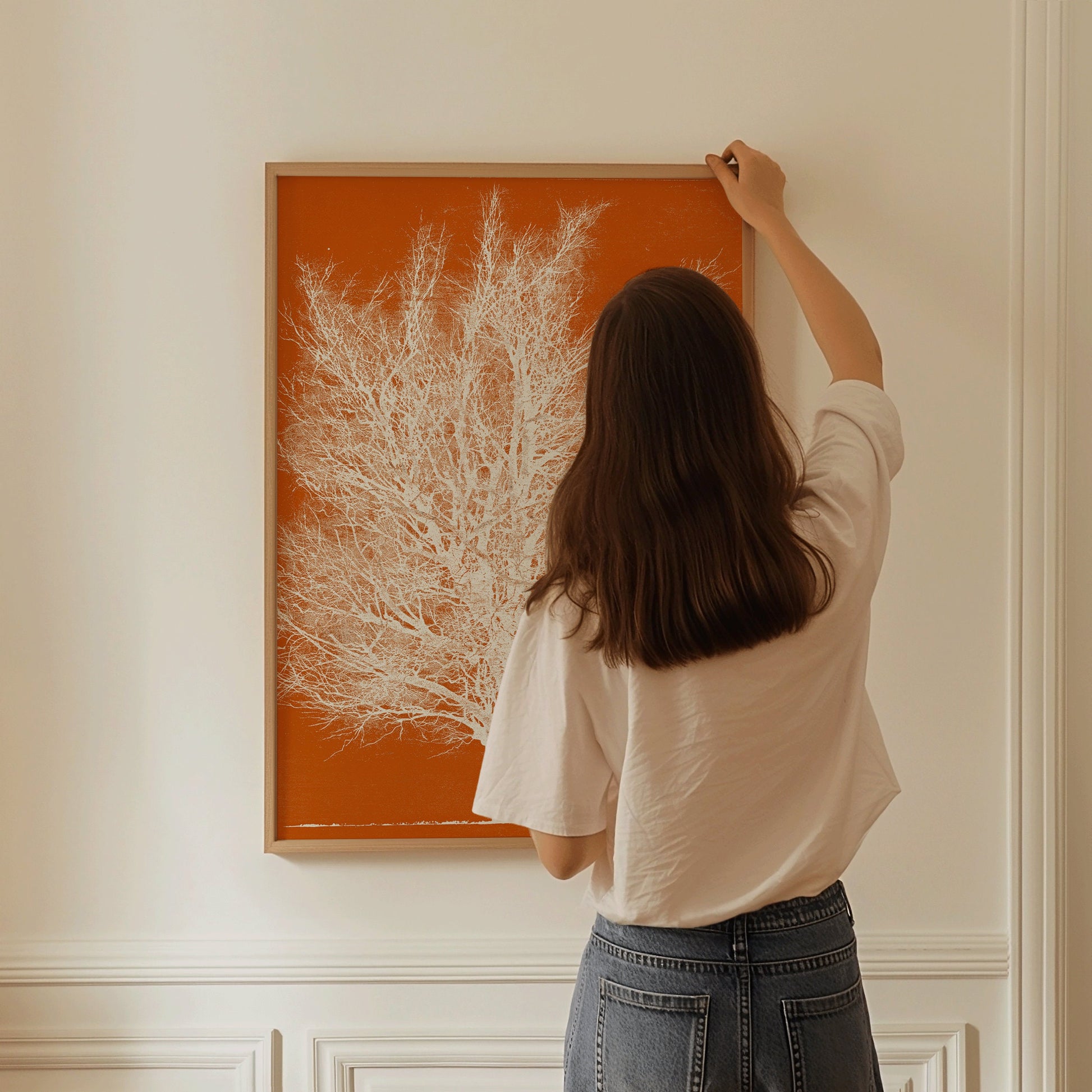 Woman hanging Joichi Hoshi's Orange and White Tree woodblock print on wall in nude oak frame.