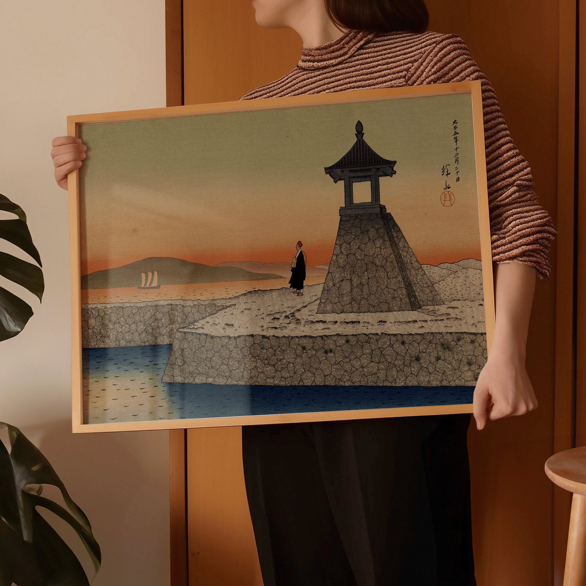 Woman holding framed Ito Shinsui Sunrise at Akashi vintage Japanese woodblock art print in a room with decorative plants.