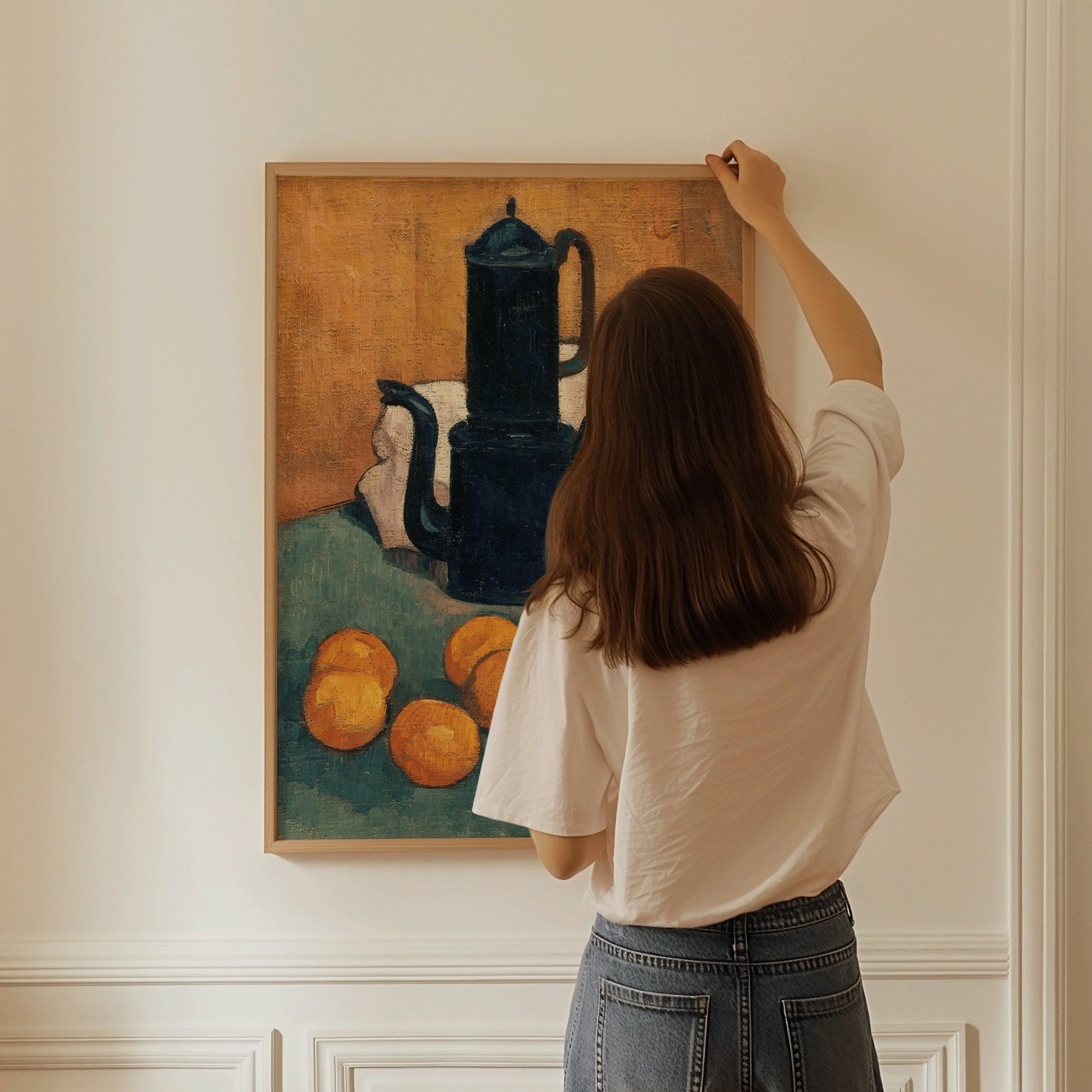 Woman hanging framed Emile Bernard's "The Blue Coffee Pot" art print on a wall, featuring impressionist still life with oranges.