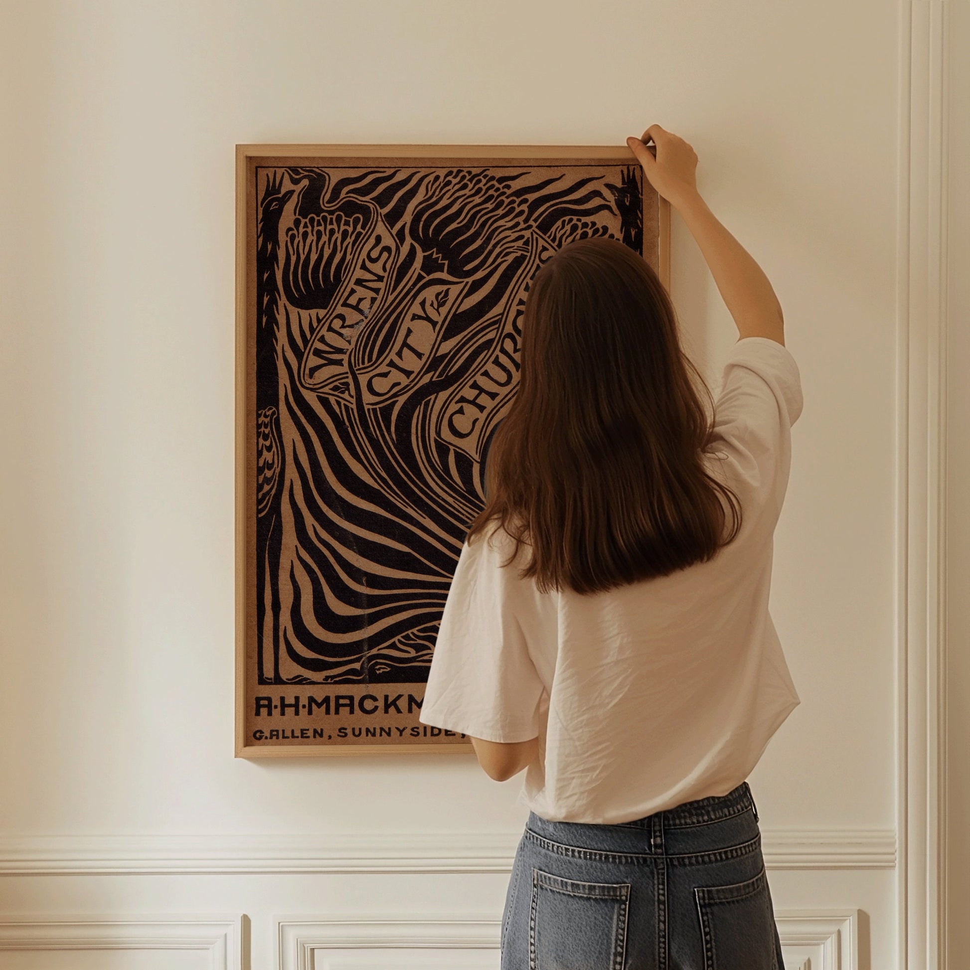 Person hanging vintage brown book cover art poster by Arthur Heygate Mackmurdo titled "Wren's City Churches" on a white wall.