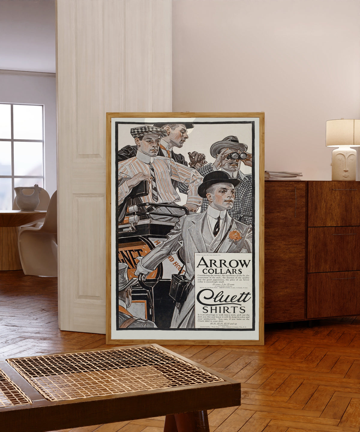 Framed art print of Arrow Collars & Cluett Shirts by J.C. Leyendecker, in a light wood frame leaning in a warm room with a parquet floor