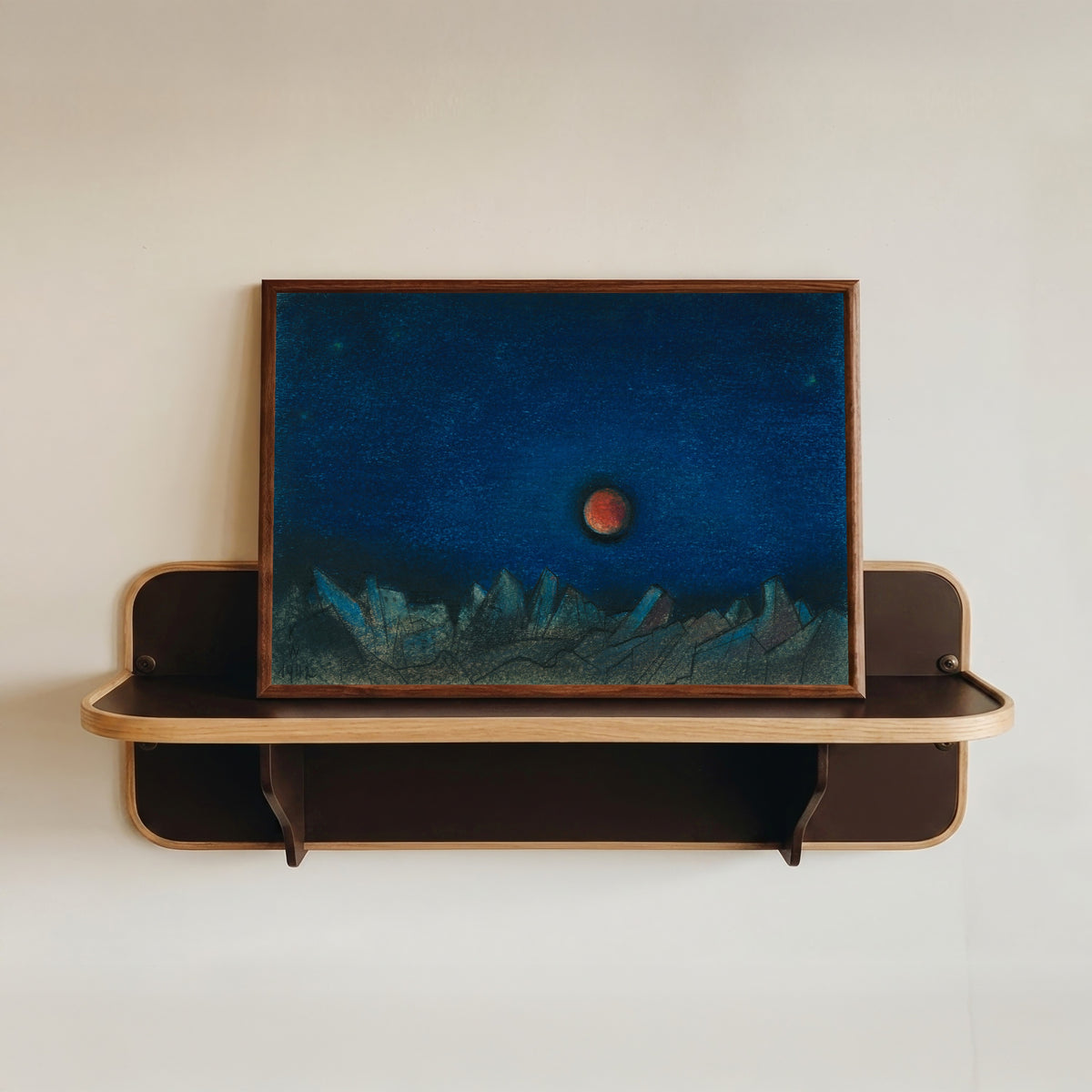 Framed art print of Untitled (Red Moon over a Landscape of Ruins) by Karl Wiener, in a dark wood frame resting on a wall shelf