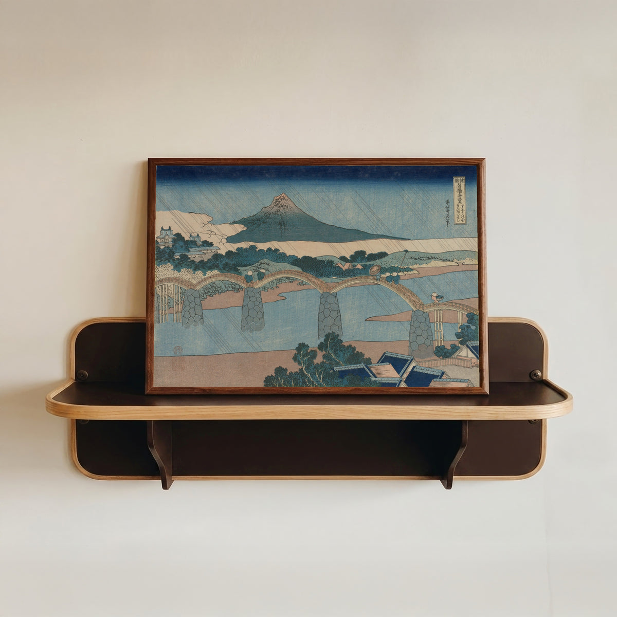Framed art print of Remarkable Views of Bridges in Various Provinces: The Brocade Bridge… by Katsushika Hokusai, in a dark wood frame resting on a wall shelf