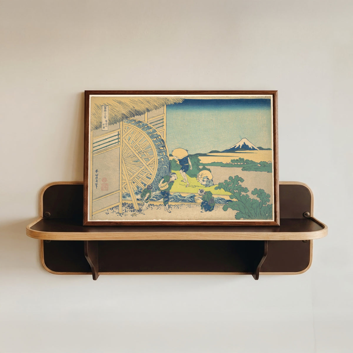 Framed art print of Thirty-six Views of Mount Fuji: Waterwheel at Onden by Katsushika Hokusai, in a dark wood frame resting on a wall shelf