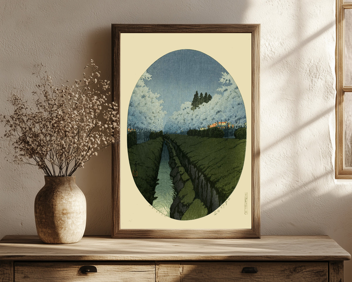 Framed art print of Cherry Blossoms at Koganei, Night by Kawase Hasui, in a dark wood frame leaning on a rustic console beside dried flowers