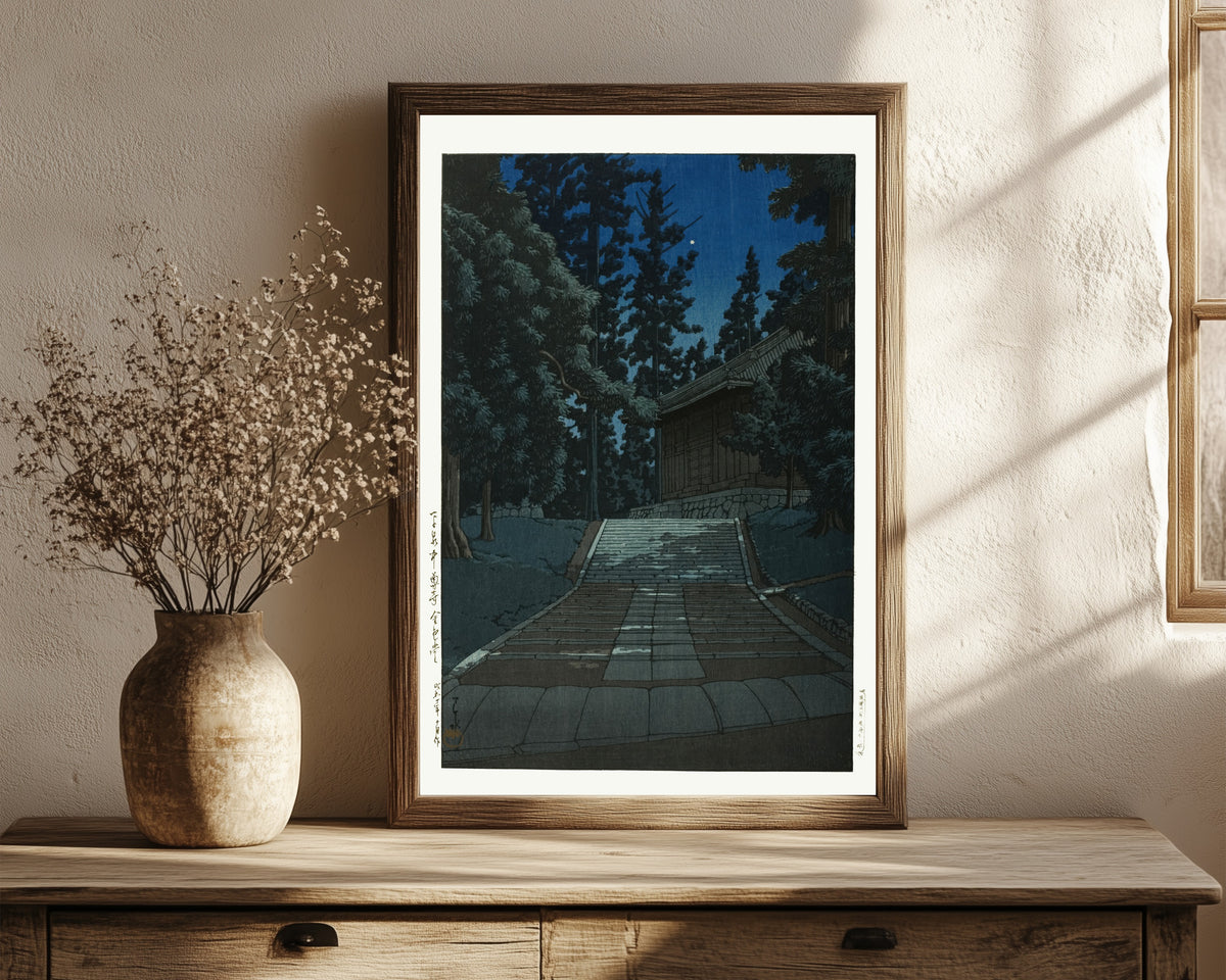 Framed art print of Konjikido at Chusonji, Hiraizumi, at Night by Kawase Hasui, in a dark wood frame leaning on a rustic console beside dried flowers