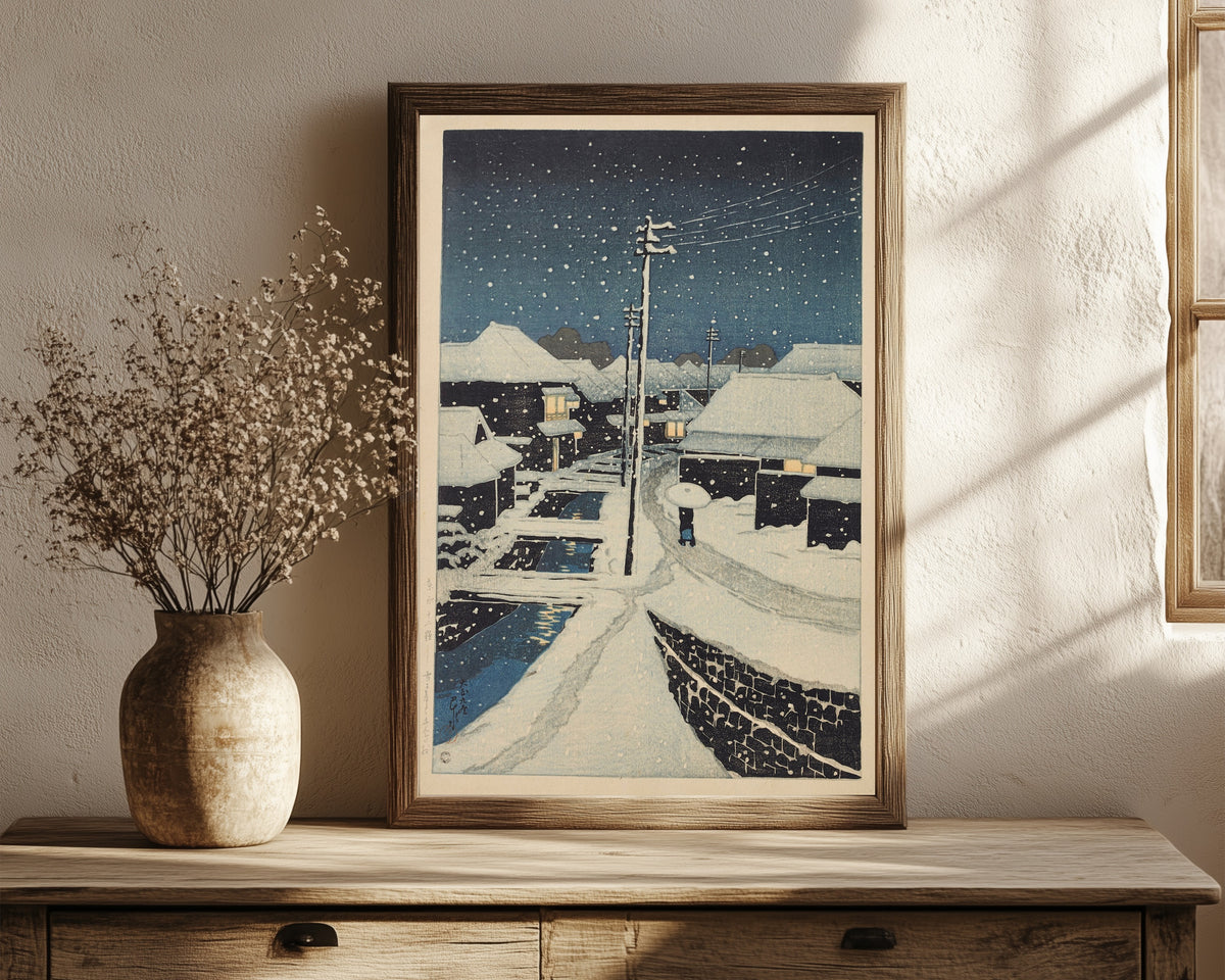 Framed art print of Evening Snow at Terajima Village by Kawase Hasui, in a dark wood frame leaning on a rustic console beside dried flowers