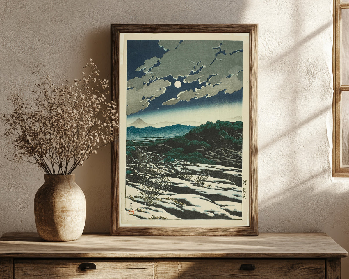 Framed art print of Karikachi Mountain Pass by Kawase Hasui, in a dark wood frame leaning on a rustic console beside dried flowers