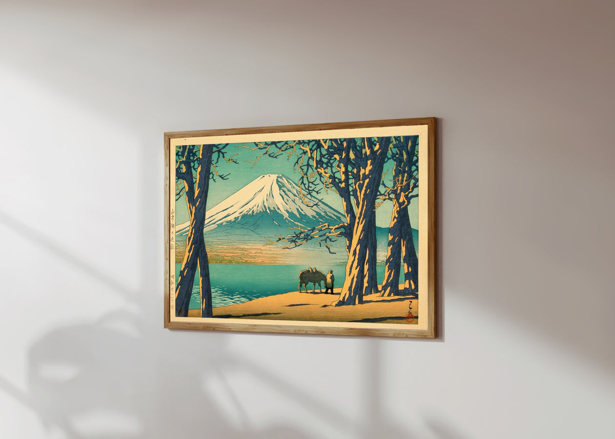Framed art print of Late Autumn at Lake Yamanaka by Kawase Hasui, in a light wood frame on a sunlit white wall, seen at a slight angle