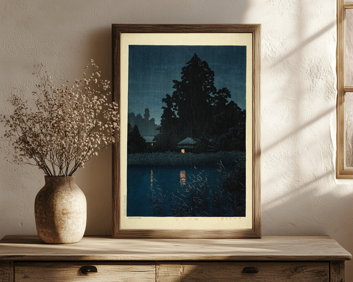 Framed art print of Omiya in the Rain by Kawase Hasui, in a dark wood frame leaning on a rustic console beside dried flowers