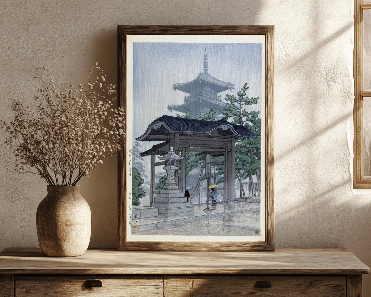Framed art print of Zentsuji Temple, Sanuki, in Rain by Kawase Hasui, in a dark wood frame leaning on a rustic console beside dried flowers