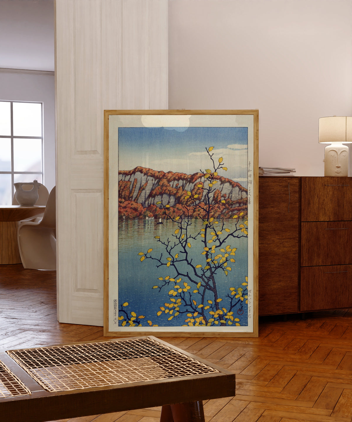 Framed art print of Lake Towada by Kawase Hasui, in a light wood frame leaning in a warm room with a parquet floor
