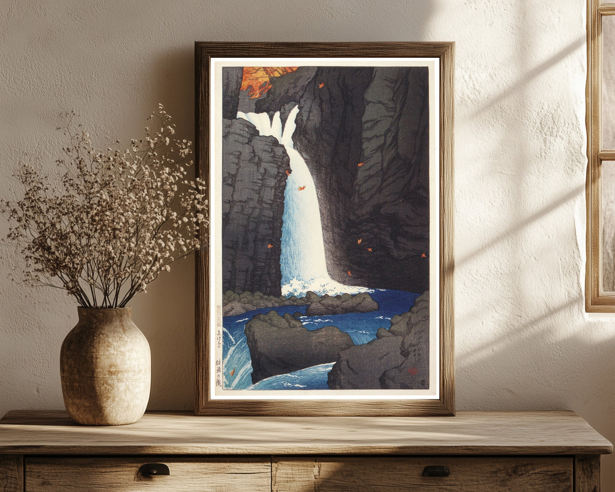 Framed art print of Shihohara Falls by Kawase Hasui, in a dark wood frame leaning on a rustic console beside dried flowers