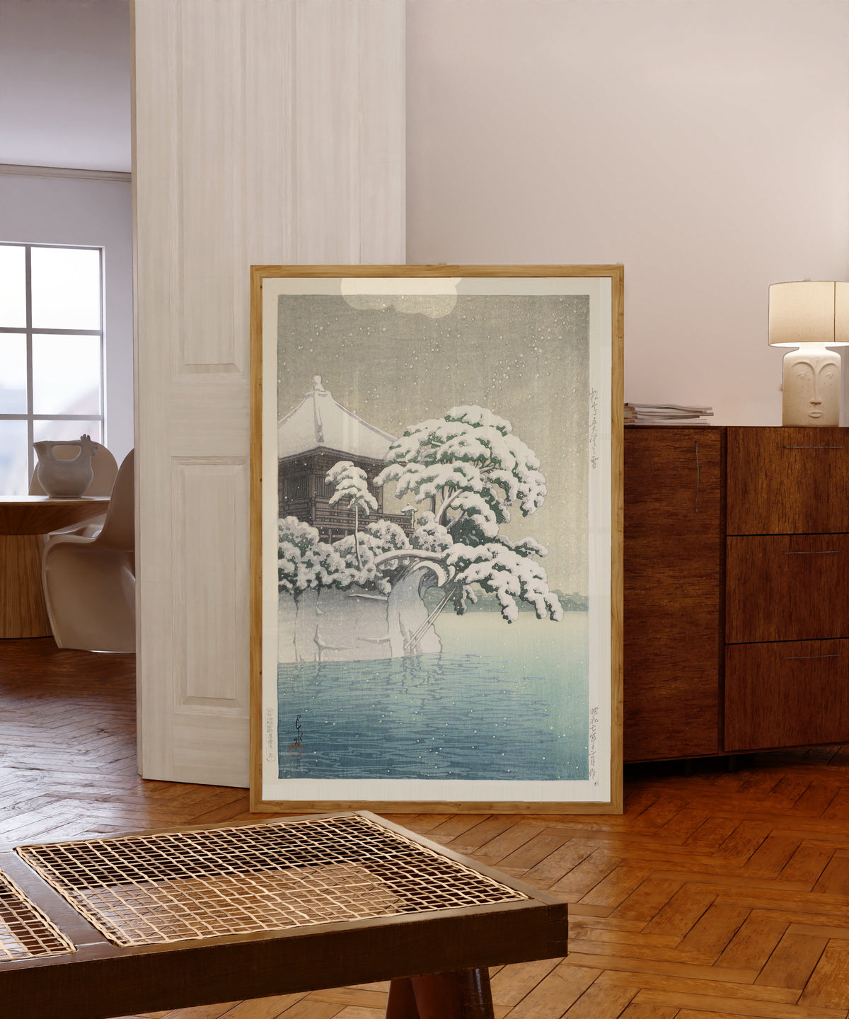 Framed art print of Snow at Godōidō, Matsushima by Kawase Hasui, in a light wood frame leaning in a warm room with a parquet floor
