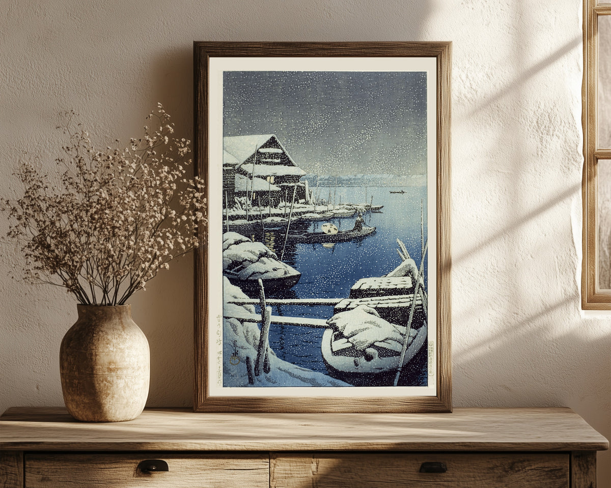 Framed art print of Snow at Mukojima by Kawase Hasui, in a dark wood frame leaning on a rustic console beside dried flowers