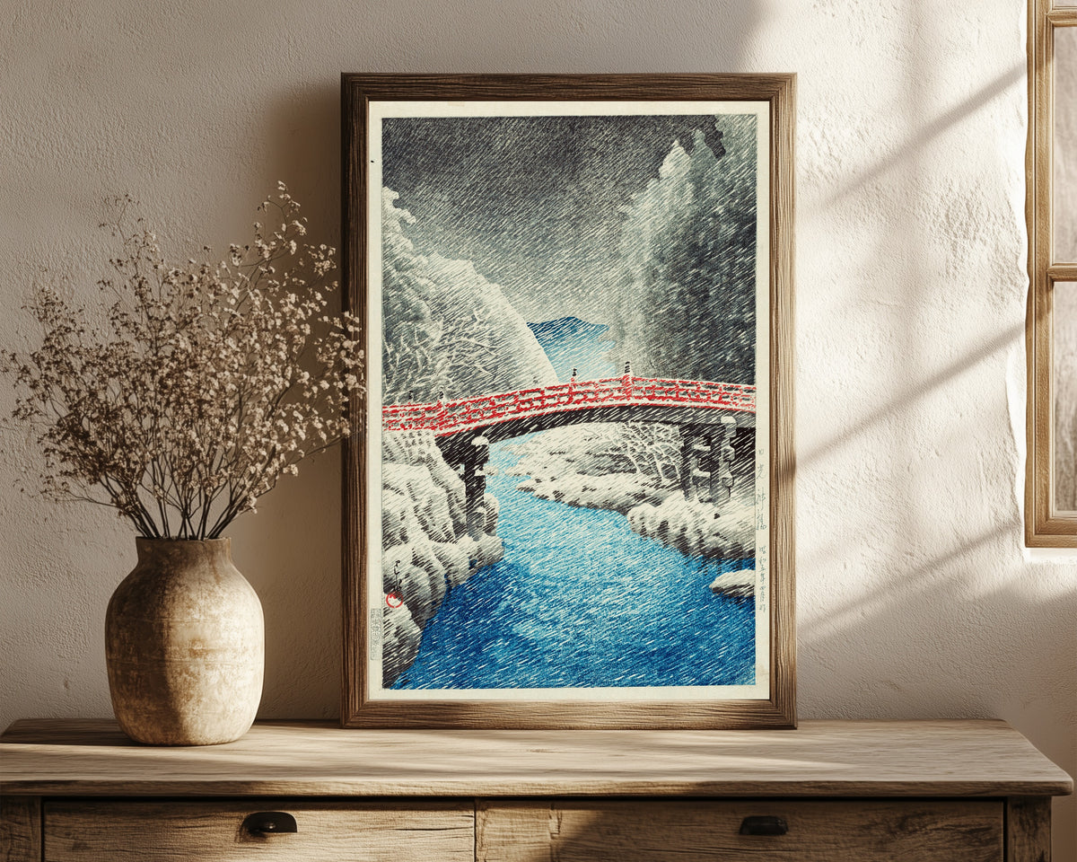 Framed art print of Snow at Shinkyo Bridge, Nikko by Kawase Hasui, in a dark wood frame leaning on a rustic console beside dried flowers
