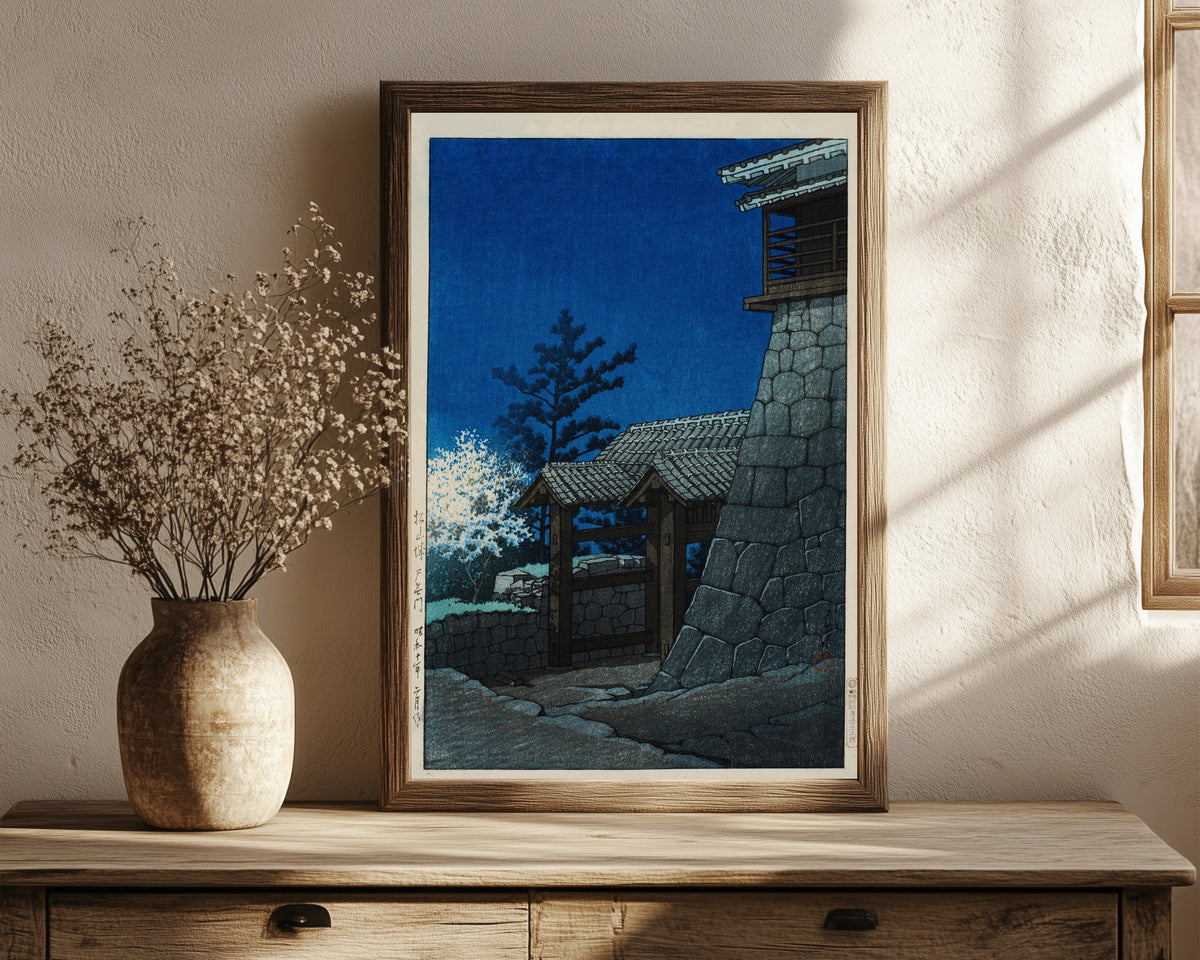 Framed art print of Tonashi Gate, Matsuyama Castle by Kawase Hasui, in a dark wood frame leaning on a rustic console beside dried flowers
