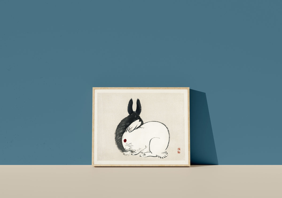 Framed art print of Black and White Rabbits by Kōno Bairei, in a light wood frame leaning on a blue wall