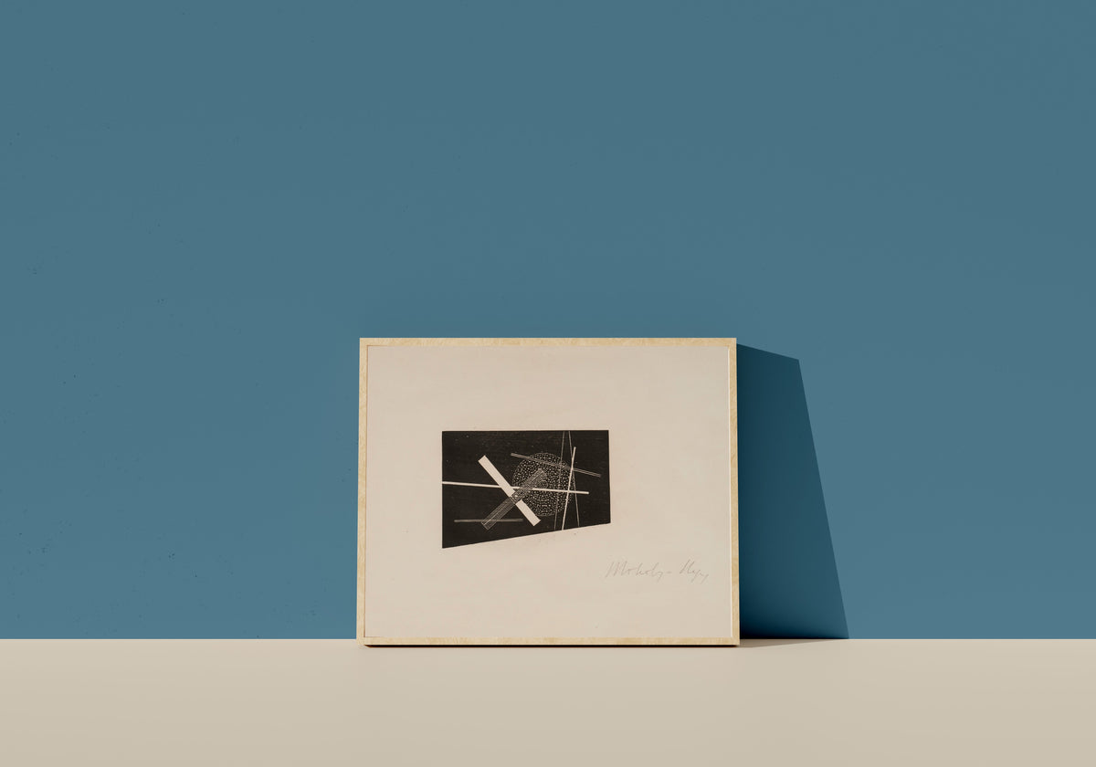 Framed art print of Composition (circa 1923) by László Moholy-Nagy, in a light wood frame leaning on a blue wall