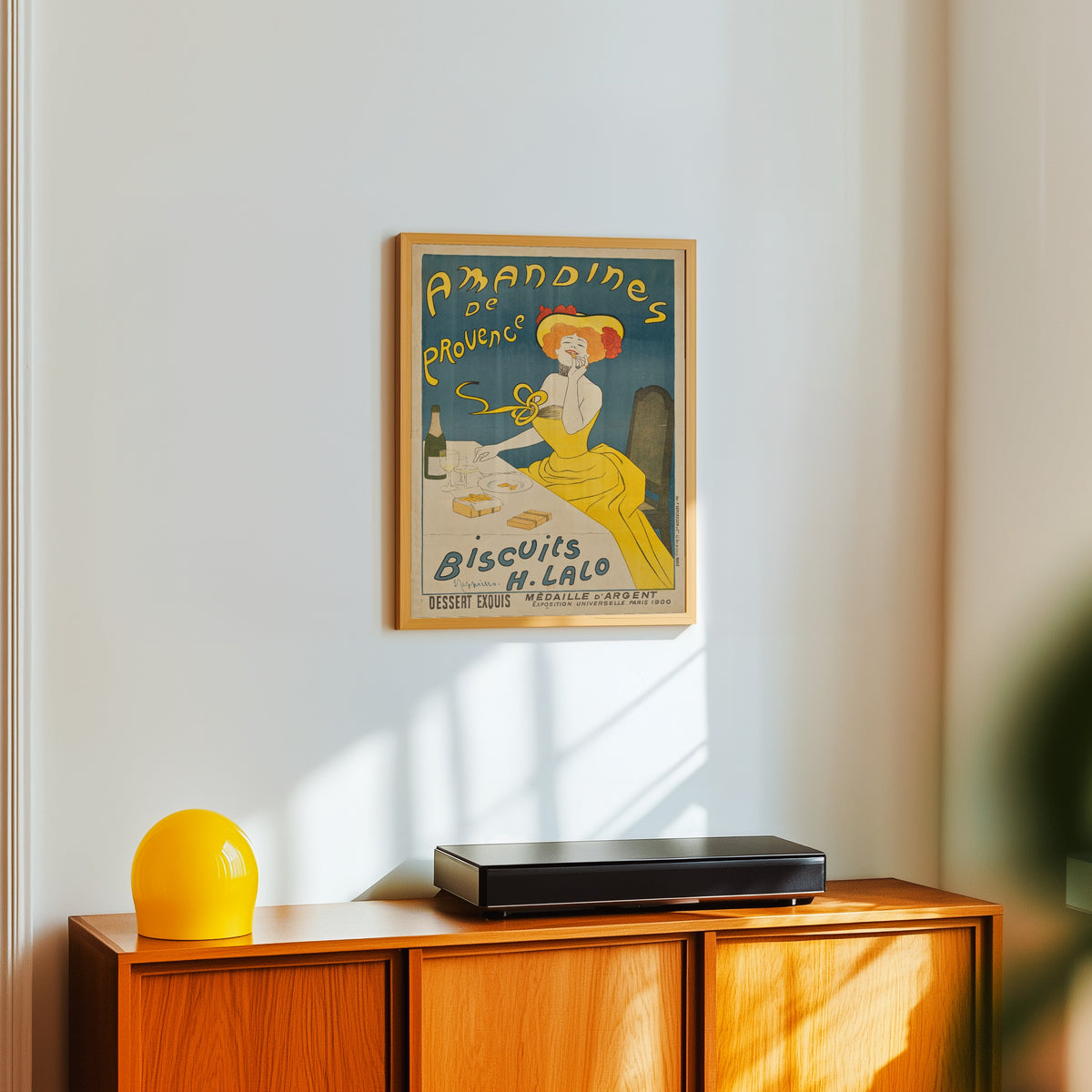 Framed art print of Amandines de Provence by Leonetto Cappiello, in a light wood frame on a white wall above an orange sideboard