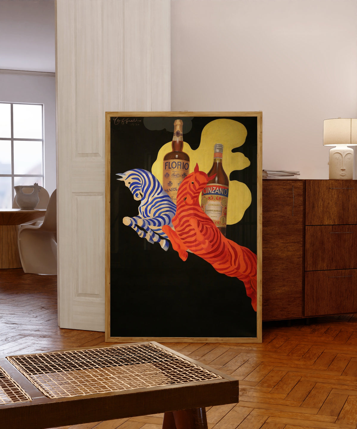 Framed art print of Florio Cinzano by Leonetto Cappiello, in a light wood frame leaning in a warm room with a parquet floor