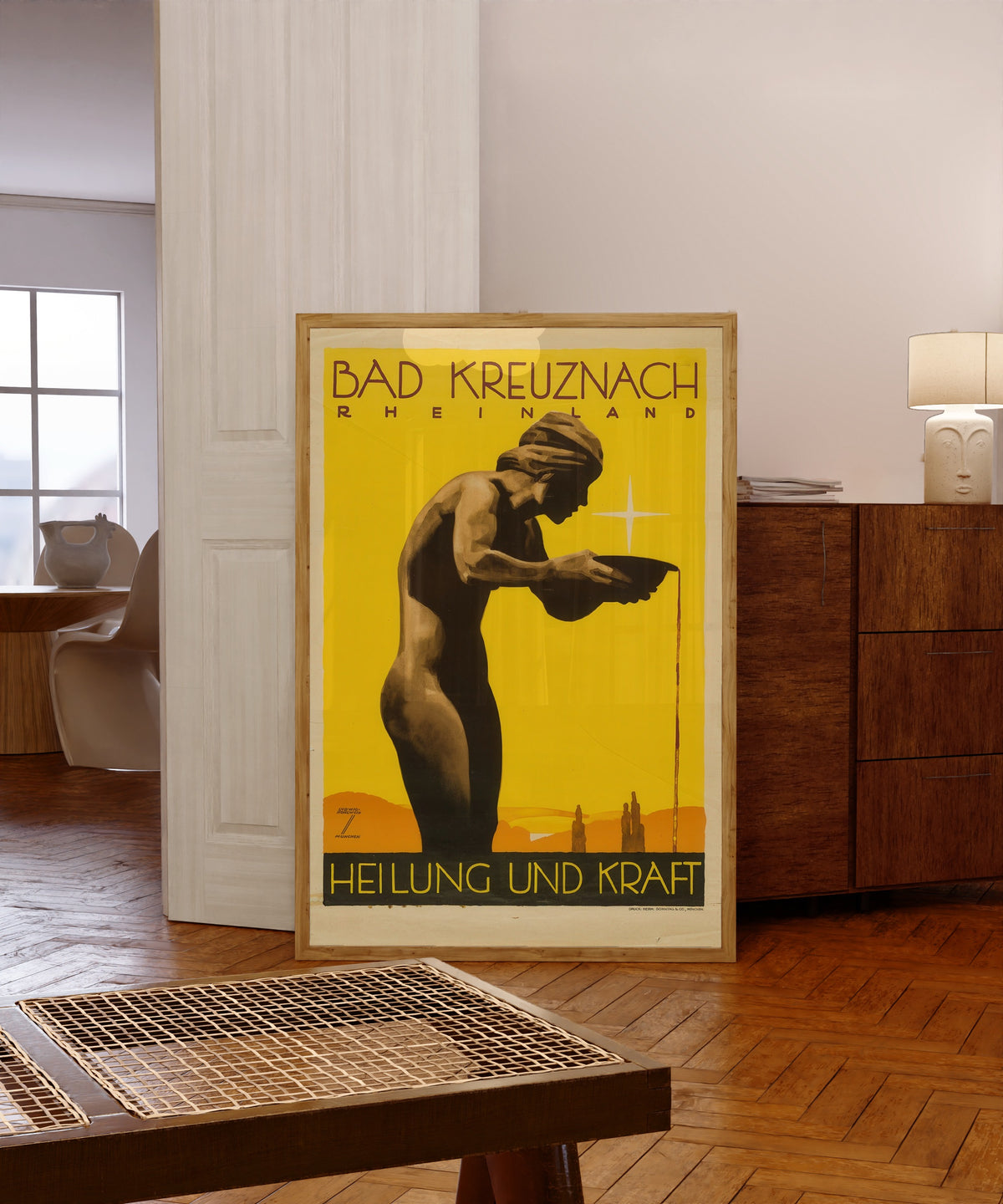 Framed art print of Bad Kreuznach by Ludwig Hohlwein, in a light wood frame leaning in a warm room with a parquet floor