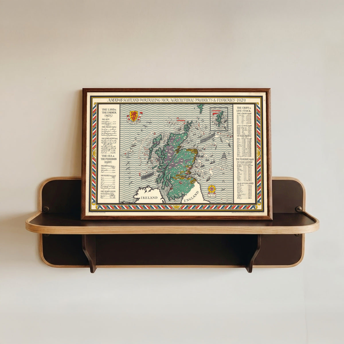 Framed art print of A Map of Scotland Portraying Her Agricultural Products and Fisheries by MacDonald Gill, in a dark wood frame resting on a wall shelf