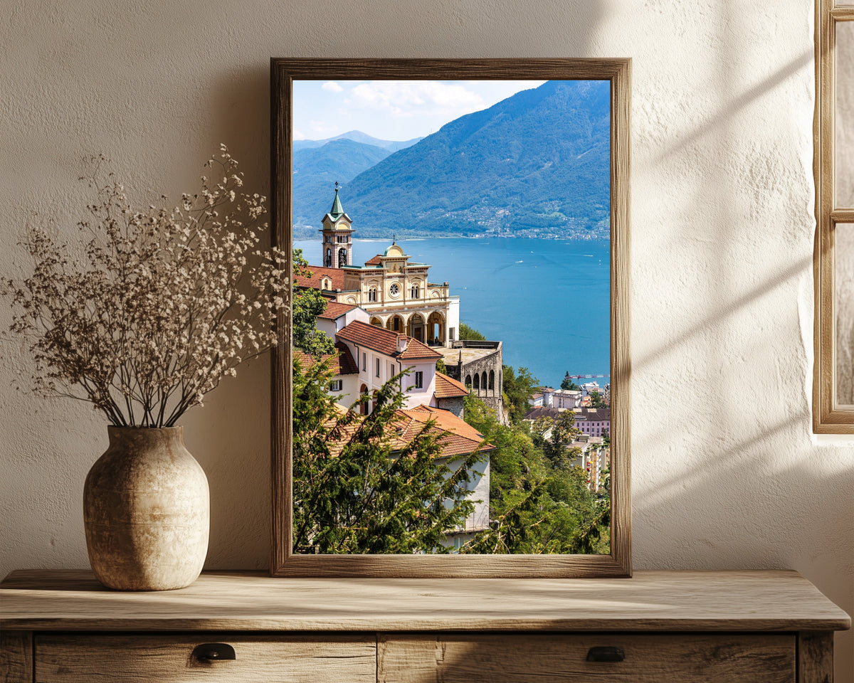 Framed art print of Madonna del Sasso in Orselina by Daniela Faust, in a dark wood frame leaning on a rustic console beside dried flowers