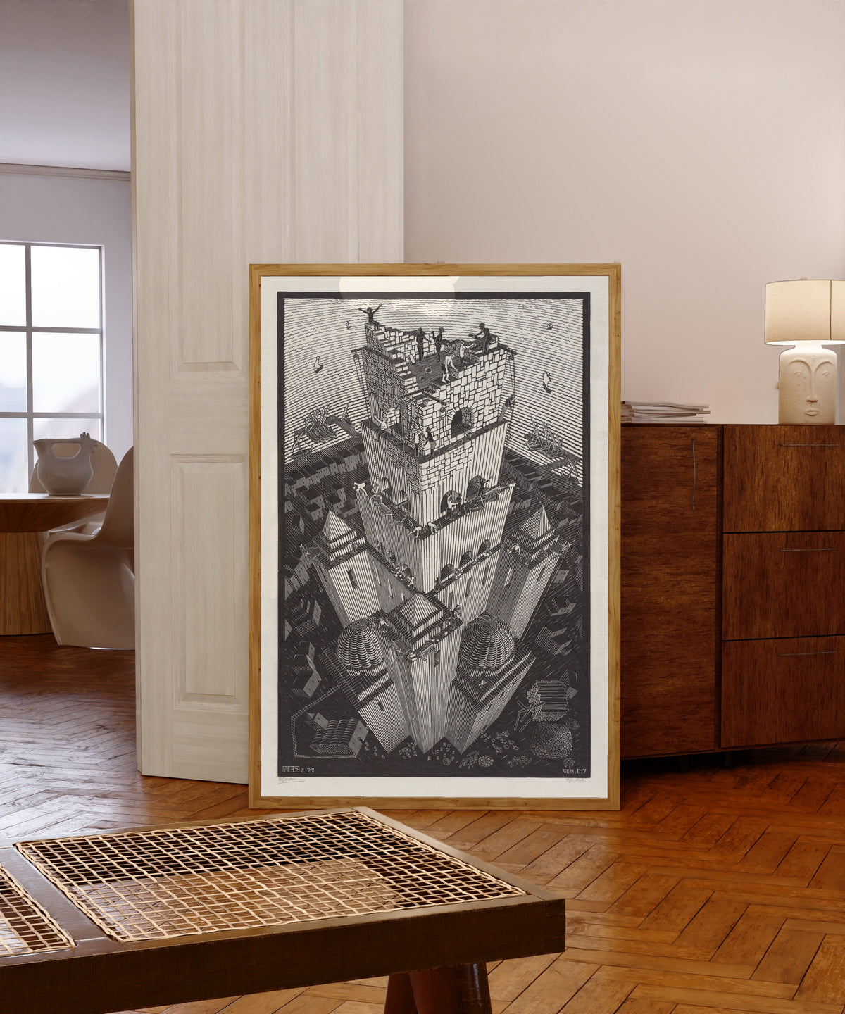 Framed art print of The Tower of Babel by M.C. Escher, in a light wood frame leaning in a warm room with a parquet floor